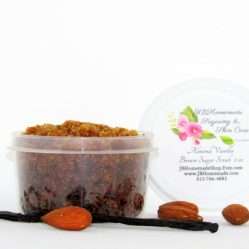 Centered is a 2 oz tub of natural brown sugar body scrub, enriched with almond, honey, and vanilla. In the foreground lies scattered almonds and a whole vanilla bean.