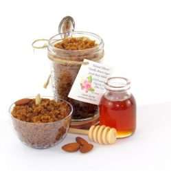 Centered is an 8 oz jar of natural brown sugar body scrub, enriched with almond, honey, and vanilla. To the right, a smaller glass jar brimming with raw honey. In the foreground lies a wooden honey dipper, surrounded by scattered almonds. To the left, a glass bowl generously filled with the sugar scrub showcases its texture, garnished with almonds on top.