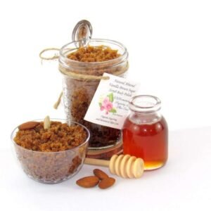 Centered is an 8 oz jar of natural brown sugar body scrub, enriched with almond, honey, and vanilla. To the right, a smaller glass jar brimming with raw honey. In the foreground lies a wooden honey dipper, surrounded by scattered almonds. To the left, a glass bowl generously filled with the sugar scrub showcases its texture, garnished with almonds on top.