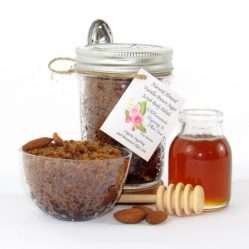 Centered is an 8 oz jar of natural brown sugar body scrub, enriched with almond, honey, and vanilla. To the right, a smaller glass jar brimming with raw honey. In the foreground lies a wooden honey dipper, surrounded by scattered almonds. To the left, a glass bowl generously filled with the sugar scrub showcases its texture, garnished with almonds on top.