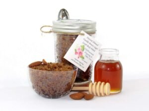 Centered is an 8 oz jar of natural brown sugar body scrub, enriched with almond, honey, and vanilla. To the right, a smaller glass jar brimming with raw honey. In the foreground lies a wooden honey dipper, surrounded by scattered almonds. To the left, a glass bowl generously filled with the sugar scrub showcases its texture, garnished with almonds on top.
