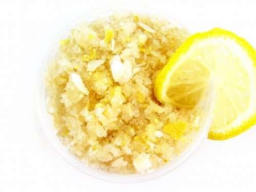A glass bowl of JBHomemade Sugaring and Skin Care's Natural Coconut Lemon Sugar Body Scrub adorned with sprinkles of dried lemon zest, coconut flakes and a lemon slice.