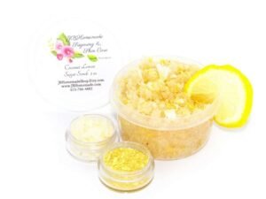 A 2 oz tub of cane sugar scrub is infused with coconut essence and zesty lemon. The sugar scrub tub is centered, topped with dried lemon zest and a slice of lemon. To the left, two tiny glass bowls filled with coconut shavings and dried lemon zest.