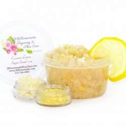 A 2 oz tub of cane sugar scrub is infused with coconut essence and zesty lemon. The sugar scrub tub is centered, topped with dried lemon zest and a slice of lemon. To the left, two tiny glass bowls filled with coconut shavings and dried lemon zest.