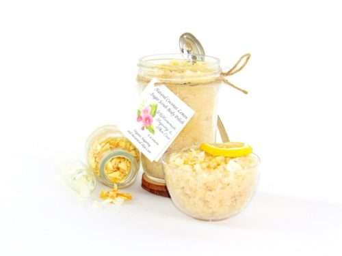 An 8 oz jar of cane sugar scrub is infused with coconut essence and zesty lemon. The sugar scrub jar is centered, topped with dried lemon zest. To the right, a glass bowl brims with the sugar scrub, adorned with a lemon slice and a sprinkle of lemon zest. On the left, a smaller glass jar lies on its side, its contents of dried lemon zest spilling onto the white surface.