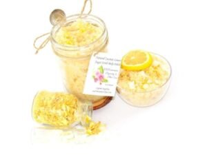 An 8 oz jar of cane sugar scrub is infused with coconut essence and zesty lemon. The sugar scrub jar is centered, topped with dried lemon zest. To the right, a glass bowl brims with the sugar scrub, adorned with a lemon slice and a sprinkle of lemon zest. On the left, a smaller glass jar lies on its side, its contents of dried lemon zest spilling onto the white surface.