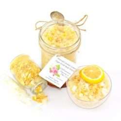 An 8 oz jar of cane sugar scrub is infused with coconut essence and zesty lemon. The sugar scrub jar is centered, topped with dried lemon zest. To the right, a glass bowl brims with the sugar scrub, adorned with a lemon slice and a sprinkle of lemon zest. On the left, a smaller glass jar lies on its side, its contents of dried lemon zest spilling onto the white surface.