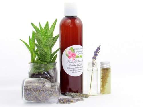 An 8 oz bottle of JBHomemade's handcrafted lavender-infused aloe vera, crafted with natural and homegrown lavender buds and aloe vera, is paired with an aloe vera plant. Alongside, a glass jar brimming with lavender buds spills over, and a small glass jar contains a sprig of lavender steeping in pure aloe vera, with two lavender sprigs resting to the right.