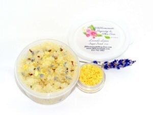 Discover a 2 oz tub of calming cane sugar scrub, a blend of relaxing lavender and invigorating lemon, ideal for a soothing experience. The sugar scrub tub is placed centrally, adorned with dried lemon zest and a lemon slice on top. Flanking the scrub tub is one smaller jar, brimming with dried lemon zest, and a delicate lavender sprig resting in front.