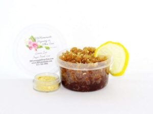 A 2 oz tub of energizing lemon zest raw and cane sugar scrub, zingy and refreshing, leaves your skin feeling invigorated. The sugar scrub tub is centered, topped with dried lemon zest and a lemon slice, with a small glass bowl filled to the brim with dried lemon zest to the left.