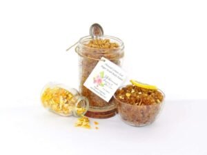 An 8 oz jar of energizing lemon zest raw and cane sugar scrub, zingy and refreshing, leaves your skin feeling invigorated. The sugar scrub jar is centered, with a glass bowl filled to the brim with the scrub to the right, topped with dried lemon zest and a lemon slice. To the left of the scrub jar lies a smaller glass jar of dried lemon zest tipped over, spilling its contents onto a white surface.
