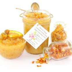 An 8 oz jar of vibrant citrus cane sugar scrub, infused with orange and calendula, is designed to enhance a radiant glow. The sugar scrub jar is placed centrally, flanked by two smaller jars—one filled with dried orange zest, the other with dried calendula—both spilling their contents onto a white backdrop. To the left, a glass bowl brims with the sugar scrub, adorned with calendula petals and crowned with a fresh orange slice.