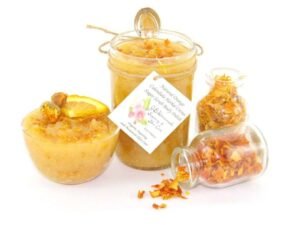 An 8 oz jar of vibrant citrus cane sugar scrub, infused with orange and calendula, is designed to enhance a radiant glow. The sugar scrub jar is placed centrally, flanked by two smaller jars—one filled with dried orange zest, the other with dried calendula—both spilling their contents onto a white backdrop. To the left, a glass bowl brims with the sugar scrub, adorned with calendula petals and crowned with a fresh orange slice.