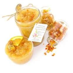 An 8 oz jar of vibrant citrus cane sugar scrub, infused with orange and calendula, is designed to enhance a radiant glow. The sugar scrub jar is placed centrally, flanked by two smaller jars—one filled with dried orange zest, the other with dried calendula—both spilling their contents onto a white backdrop. To the left, a glass bowl brims with the sugar scrub, adorned with calendula petals and crowned with a fresh orange slice.