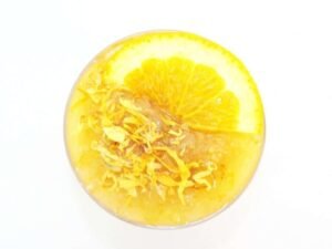 A glass bowl of JBHomemade Sugaring and Skin Care's Natural Orange Calendula Herbal Citrus Sugar Body Scrub adorned with sprinkles of dried calendula and an orange slice.