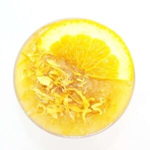 A glass bowl of JBHomemade Sugaring and Skin Care's Natural Orange Calendula Herbal Citrus Sugar Body Scrub adorned with sprinkles of dried calendula and an orange slice.