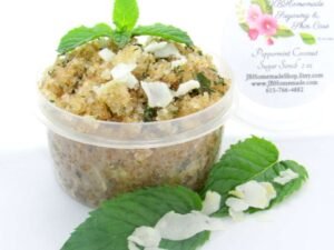 A 2 oz tub of cooling peppermint and coconut cane sugar scrub offers a refreshing and hydrating experience for your skin. The sugar scrub tub is centrally placed, flanked by bright green sprigs of peppermint and coconut flakes are scattered throughout the arrangement.