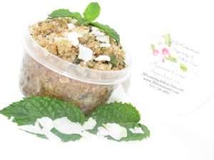 A 2 oz tub of cooling peppermint and coconut cane sugar scrub offers a refreshing and hydrating experience for your skin. The sugar scrub tub is centrally placed, flanked by bright green sprigs of peppermint and coconut flakes are scattered throughout the arrangement.