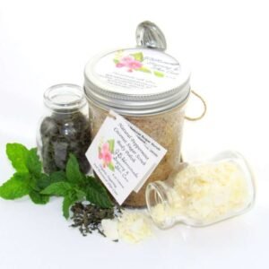 An 8 oz jar of cooling peppermint and coconut cane sugar scrub offers a refreshing and hydrating experience for your skin. The sugar scrub jar is centrally placed, flanked by two smaller glass jars—one holding dried peppermint, the other coconut flakes—with their contents artfully strewn around. Bright green sprigs of peppermint are scattered throughout the arrangement.