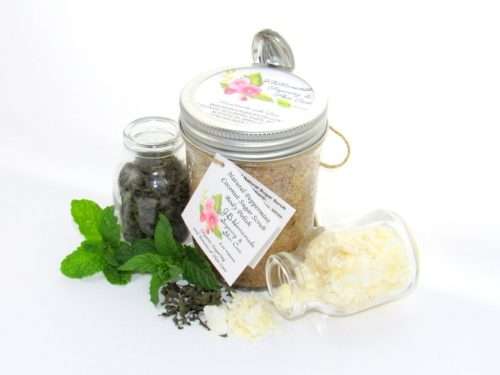 An 8 oz jar of cooling peppermint and coconut cane sugar scrub offers a refreshing and hydrating experience for your skin. The sugar scrub jar is centrally placed, flanked by two smaller glass jars—one holding dried peppermint, the other coconut flakes—with their contents artfully strewn around. Bright green sprigs of peppermint are scattered throughout the arrangement.