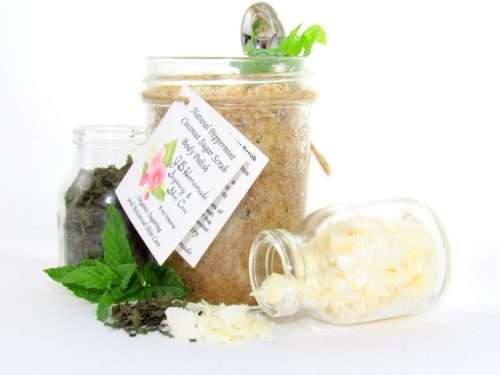 An 8 oz jar of cooling peppermint and coconut cane sugar scrub offers a refreshing and hydrating experience for your skin. The sugar scrub jar is centrally placed, flanked by two smaller glass jars—one holding dried peppermint, the other coconut flakes—with their contents artfully strewn around. Bright green sprigs of peppermint are scattered throughout the arrangement.