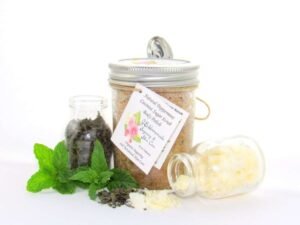 An 8 oz jar of cooling peppermint and coconut cane sugar scrub offers a refreshing and hydrating experience for your skin. The sugar scrub jar is centrally placed, flanked by two smaller glass jars—one holding dried peppermint, the other coconut flakes—with their contents artfully strewn around. Bright green sprigs of peppermint are scattered throughout the arrangement.