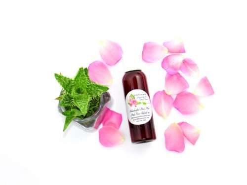 An overhead perspective of a 2 oz bottle of handcrafted Radiant Rose Glow: Pure Aloe Vera Infused with Pink Rose Petals with natural ingredients of pink rose petals and an aloe vera plant visible.