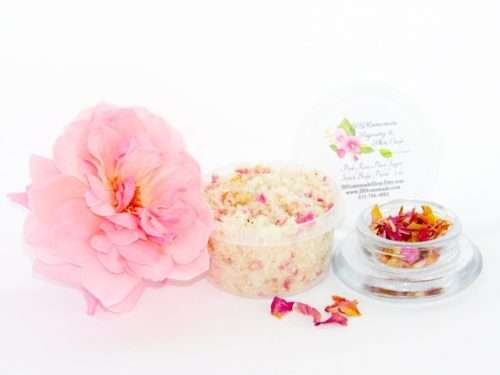 Explore the delicate 2 oz tub of soft pink rose petal cane sugar scrub, gently scented and perfect for pampering your skin. To the left, the tub is placed next to a glass bowl filled with dried pink rose petals. A tender pink rose bloom rests beside the sugar scrub, with dried pink rose petals scattered around.