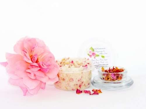 Explore the delicate 2 oz tub of soft pink rose petal cane sugar scrub, gently scented and perfect for pampering your skin. To the left, the tub is placed next to a glass bowl filled with dried pink rose petals. A tender pink rose bloom rests beside the sugar scrub, with dried pink rose petals scattered around.