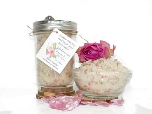 Discover an 8 oz jar of soft pink rose petal cane sugar scrub, subtly fragranced and ideal for treating your skin. To the left, the jar is positioned beside a glass bowl brimming with the scrub, crowned with a vivid pink rose. Surrounding it, pink rose petals and beads are artfully strewn.
