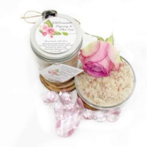 Discover an 8 oz jar of soft pink rose petal cane sugar scrub, subtly fragranced and ideal for treating your skin. To the left, the jar is positioned beside a glass bowl brimming with the scrub, crowned with a vivid pink rose. Surrounding it, pink rose petals and beads are artfully strewn.