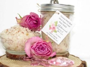 Discover an 8 oz jar of soft pink rose petal cane sugar scrub, subtly fragranced and ideal for treating your skin. To the left, the jar is positioned beside a glass bowl brimming with the scrub, crowned with a vivid pink rose. Surrounding it, pink rose petals and beads are artfully strewn.