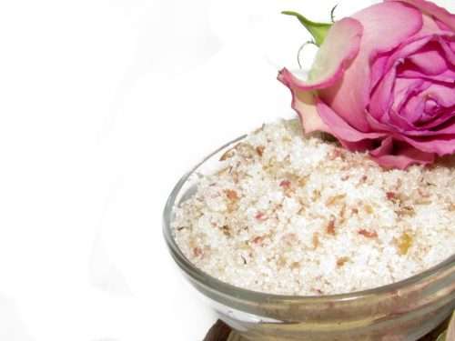 A glass bowl of JBHomemade Sugaring and Skin Care's Natural Pink Rose Petal Floral Sugar Body Scrub with a fresh pink rose bloom sitting on top.