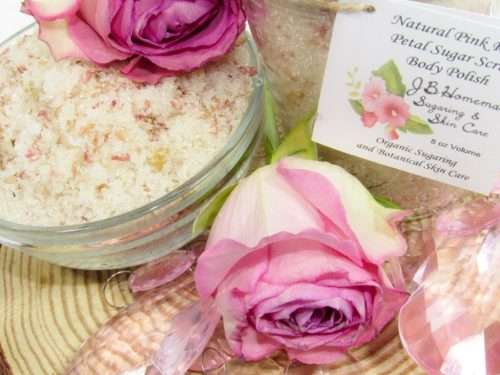Discover an 8 oz jar of soft pink rose petal cane sugar scrub, subtly fragranced and ideal for treating your skin. To the left, the jar is positioned beside a glass bowl brimming with the scrub, crowned with a vivid pink rose. Surrounding it, pink rose petals and beads are artfully strewn.