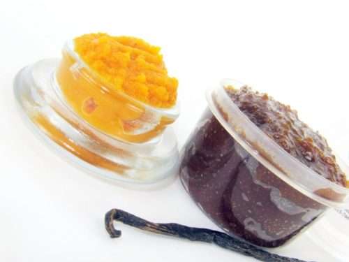Indulge in the seasonal warmth of a 2 oz tub of pumpkin vanilla brown sugar scrub. The jar is prominently displayed at the center, accompanied by a glass bowl showcasing the scrub's rich texture. To the left, a smaller bowl contains pure pumpkin puree and a whole vanilla bean, highlighting the scrub's natural ingredients.