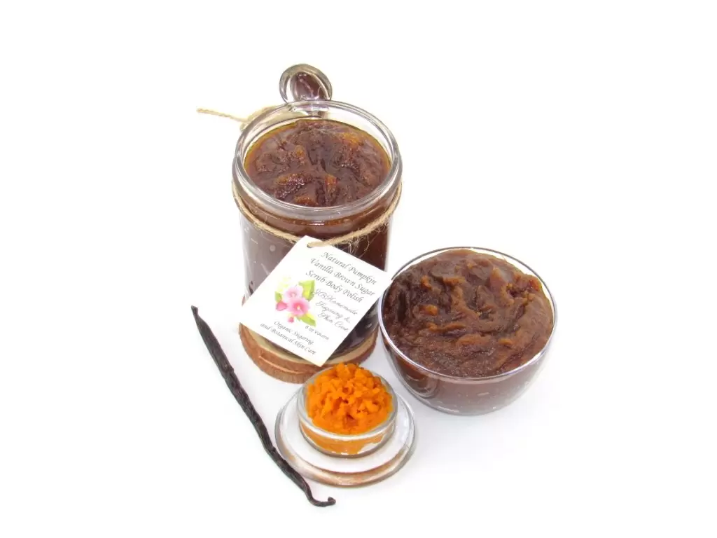 Indulge in the seasonal warmth of an 8 oz jar of pumpkin vanilla brown sugar scrub. The jar is prominently displayed at the center, accompanied by a glass bowl showcasing the scrub's rich texture. To the left, a smaller bowl contains pure pumpkin puree and a whole vanilla bean, highlighting the scrub's natural ingredients.