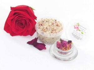Discover a 2 oz tub of exfoliating cane sugar scrub, enriched with aromatic red rose petals, offers a calming and opulent skin treatment. The scrub's tub is centrally placed, with a glass bowl to the right filled with the sugar scrub and crowned by a vivid, fully bloomed red rose. In the foreground, a smaller glass bowl contains dried rose petals, with additional petals strewn across a white backdrop.