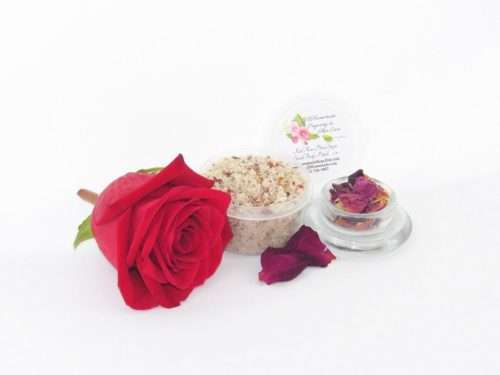 Discover a 2 oz tub of exfoliating cane sugar scrub, enriched with aromatic red rose petals, offers a calming and opulent skin treatment. The scrub's tub is centrally placed, with a glass bowl to the right filled with the sugar scrub and crowned by a vivid, fully bloomed red rose. In the foreground, a smaller glass bowl contains dried rose petals, with additional petals strewn across a white backdrop.