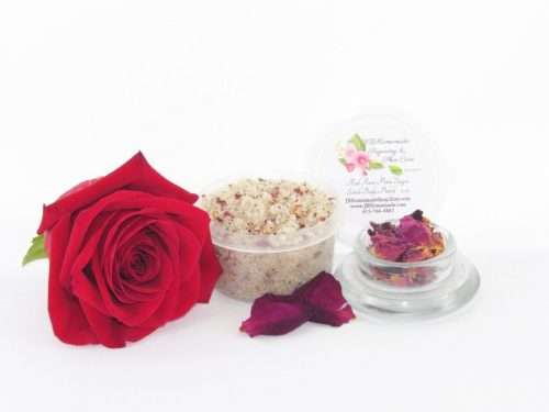 Discover a 2 oz tub of exfoliating cane sugar scrub, enriched with aromatic red rose petals, offers a calming and opulent skin treatment. The scrub's tub is centrally placed, with a glass bowl to the right filled with the sugar scrub and crowned by a vivid, fully bloomed red rose. In the foreground, a smaller glass bowl contains dried rose petals, with additional petals strewn across a white backdrop.
