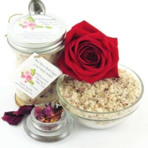 An 8 oz jar of exfoliating cane sugar scrub, enriched with aromatic red rose petals, offers a calming and opulent skin treatment. The scrub's jar is centrally placed, with a glass bowl to the right filled with the sugar scrub and crowned by a vivid, fully bloomed red rose. In the foreground, a smaller glass bowl contains dried rose petals, with additional petals strewn across a white backdrop.