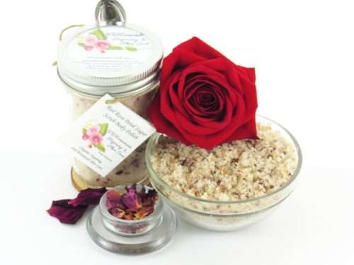 An 8 oz jar of exfoliating cane sugar scrub, enriched with aromatic red rose petals, offers a calming and opulent skin treatment. The scrub's jar is centrally placed, with a glass bowl to the right filled with the sugar scrub and crowned by a vivid, fully bloomed red rose. In the foreground, a smaller glass bowl contains dried rose petals, with additional petals strewn across a white backdrop.