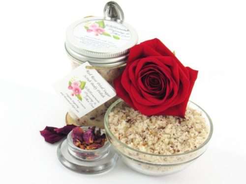 An 8 oz jar of exfoliating cane sugar scrub, enriched with aromatic red rose petals, offers a calming and opulent skin treatment. The scrub's jar is centrally placed, with a glass bowl to the right filled with the sugar scrub and crowned by a vivid, fully bloomed red rose. In the foreground, a smaller glass bowl contains dried rose petals, with additional petals strewn across a white backdrop.