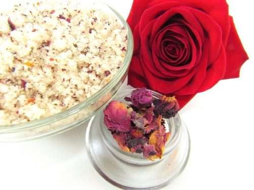 Discover a glass bowl of exfoliating cane sugar scrub, enriched with aromatic red rose petals, offers a calming and opulent skin treatment. The scrub is crowned by a vivid, fully bloomed red rose. In the foreground, a smaller glass bowl contains dried rose petals.