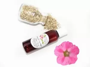 A bottle of JBHomemade’s handcrafted hollyhock water toner, made with organic marshmallow root and fresh hollyhock blooms, accompanied by dried marshmallow root and a vibrant hollyhock flower.