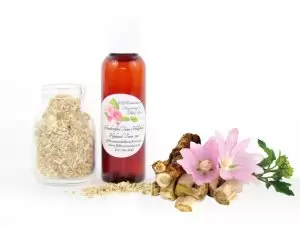 A bottle of JBHomemade’s handcrafted hollyhock water toner, made with organic marshmallow root and fresh hollyhock blooms, accompanied by dried marshmallow root and a vibrant hollyhock flower.