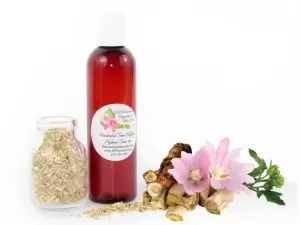 A bottle of JBHomemade’s handcrafted hollyhock water toner, made with organic marshmallow root and fresh hollyhock blooms, accompanied by dried marshmallow root and a vibrant hollyhock flower.