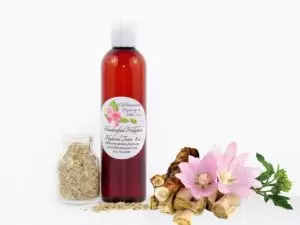 A bottle of JBHomemade’s handcrafted hollyhock water toner, made with organic marshmallow root and fresh hollyhock blooms, accompanied by dried marshmallow root and a vibrant hollyhock flower.