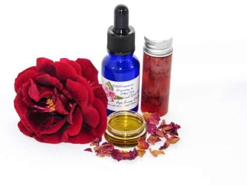 A bottle of ‘Rose the Age Away Natural Facial Serum’ surrounded by its natural ingredients, avocado oil, rose oil, and rose petals.