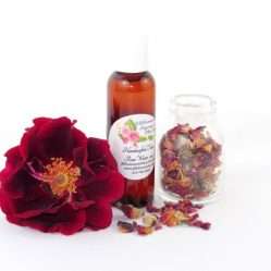 A 2 oz bottle of handcrafted rose water is presented with a vibrant red rose to the left and dried rose petals scattered from a glass jar on the right, capturing the pure and organic quality of JBHomemade's garden-fresh rose water.