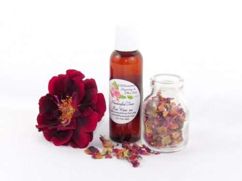 A 2 oz bottle of handcrafted rose water is presented with a vibrant red rose to the left and dried rose petals scattered from a glass jar on the right, capturing the pure and organic quality of JBHomemade's garden-fresh rose water.